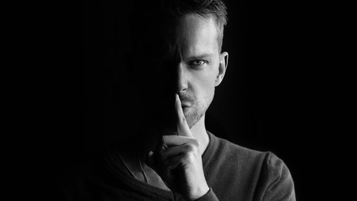 Black and white portrait of a man gesturing for silence, highlighting mystery.